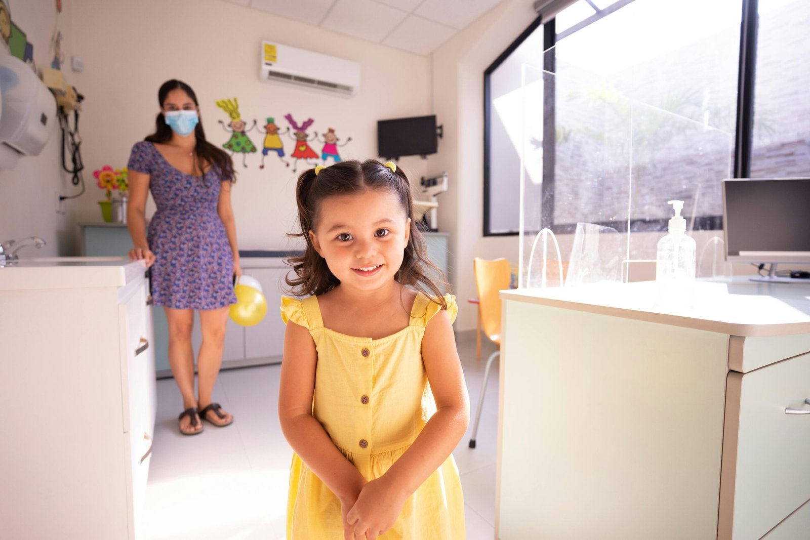 A cheerful child in a yellow dress at a pediatric clinic with doctor in background.